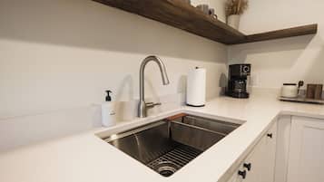A modern kitchen with a stainless steel sink, white countertops, wooden shelves, and a coffee maker.