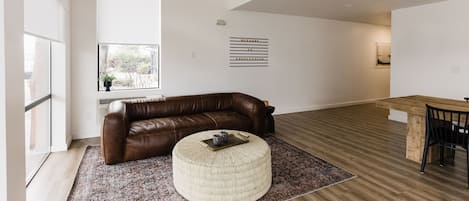 A spacious living area with a brown leather sofa, a round ottoman, and a wooden dining table.