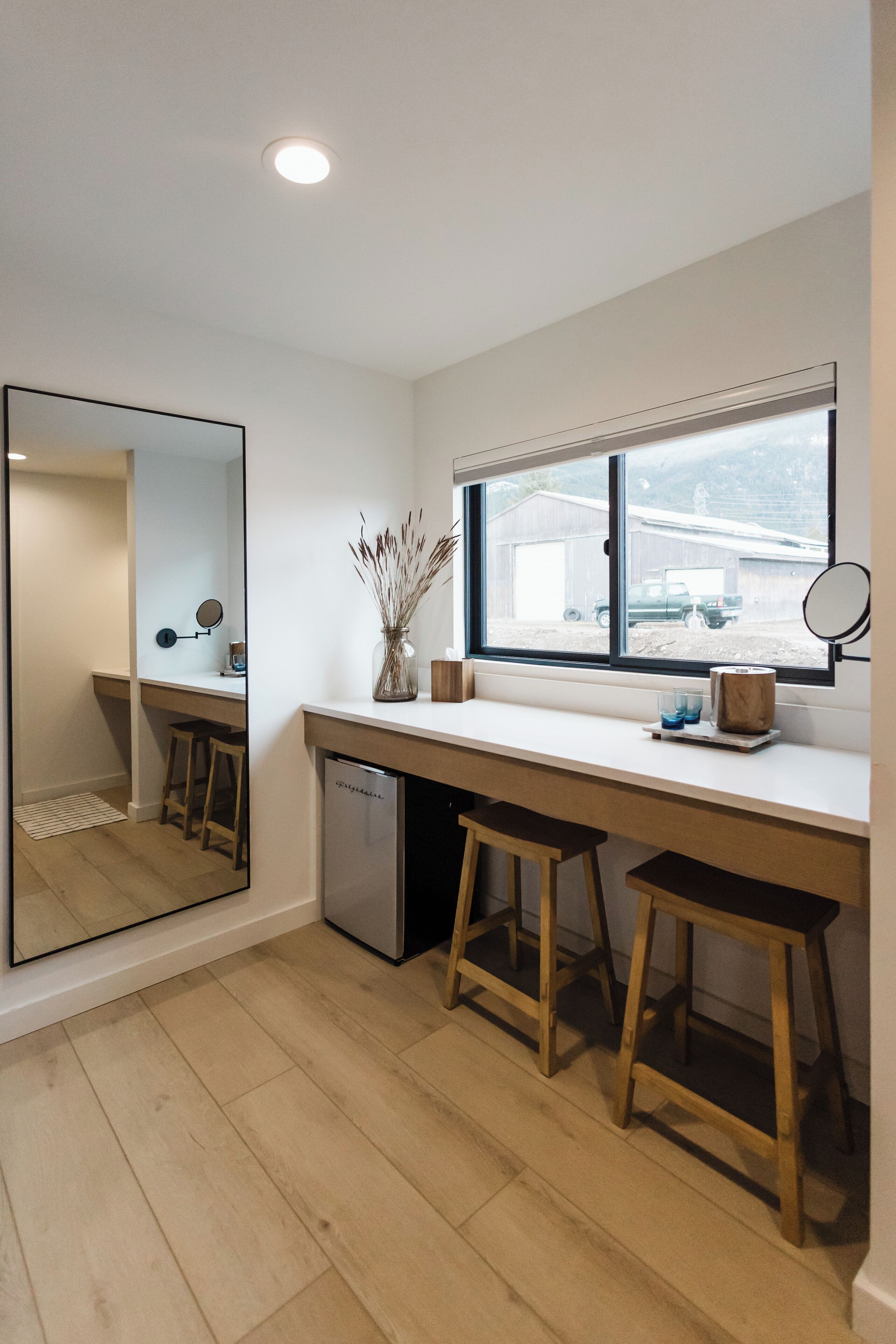 A compact kitchen with a white countertop, wooden stools, a small sink, and a window with a view of a building outside.