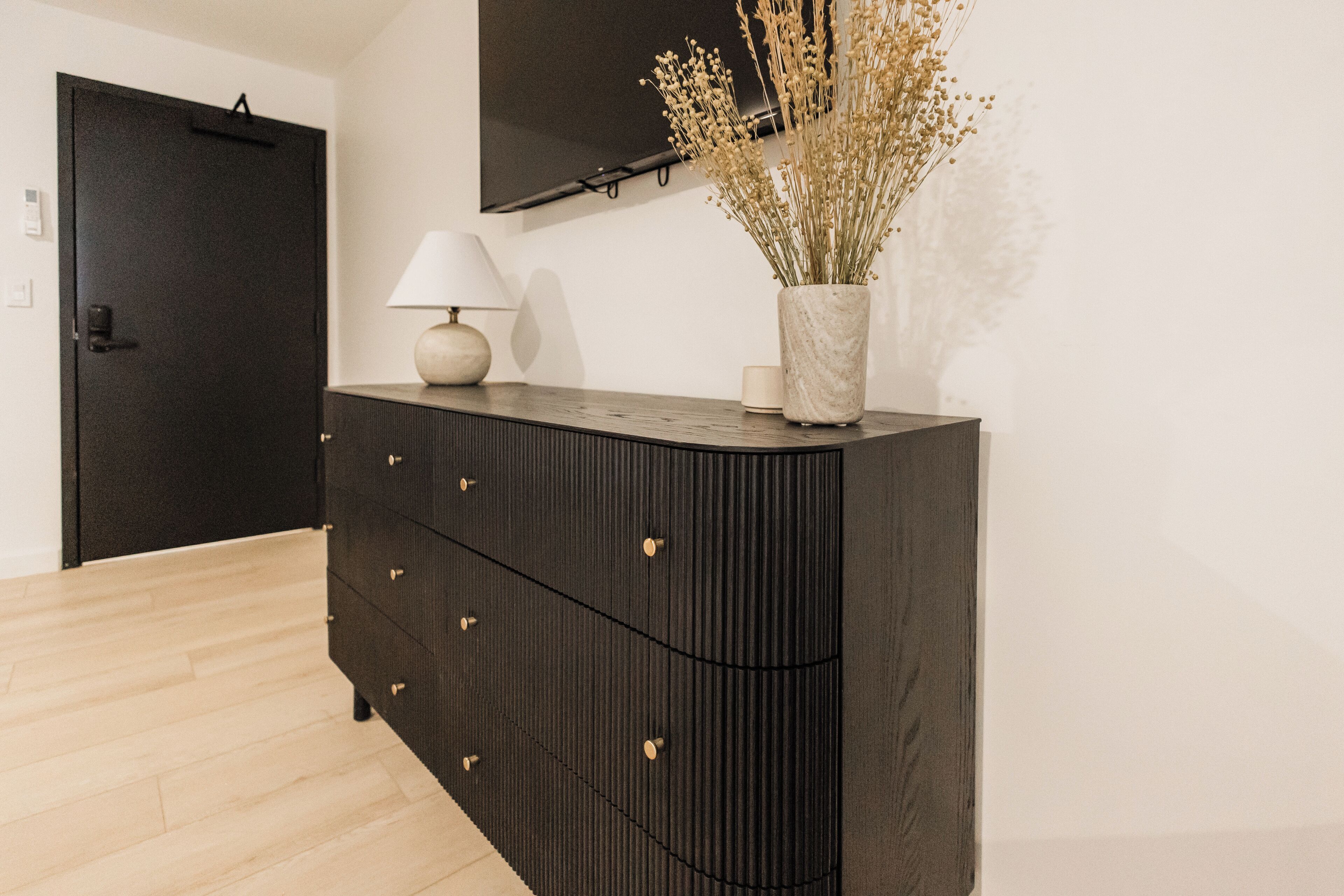 A modern room with a dark wooden dresser, a flat-screen TV mounted on the wall, and a vase with dried flowers on the dresser.