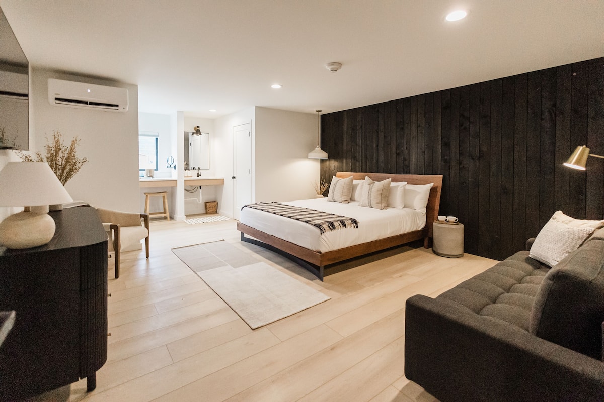 A modern bedroom with a large bed, a dark wooden accent wall, a bedside lamp, and a small round table.