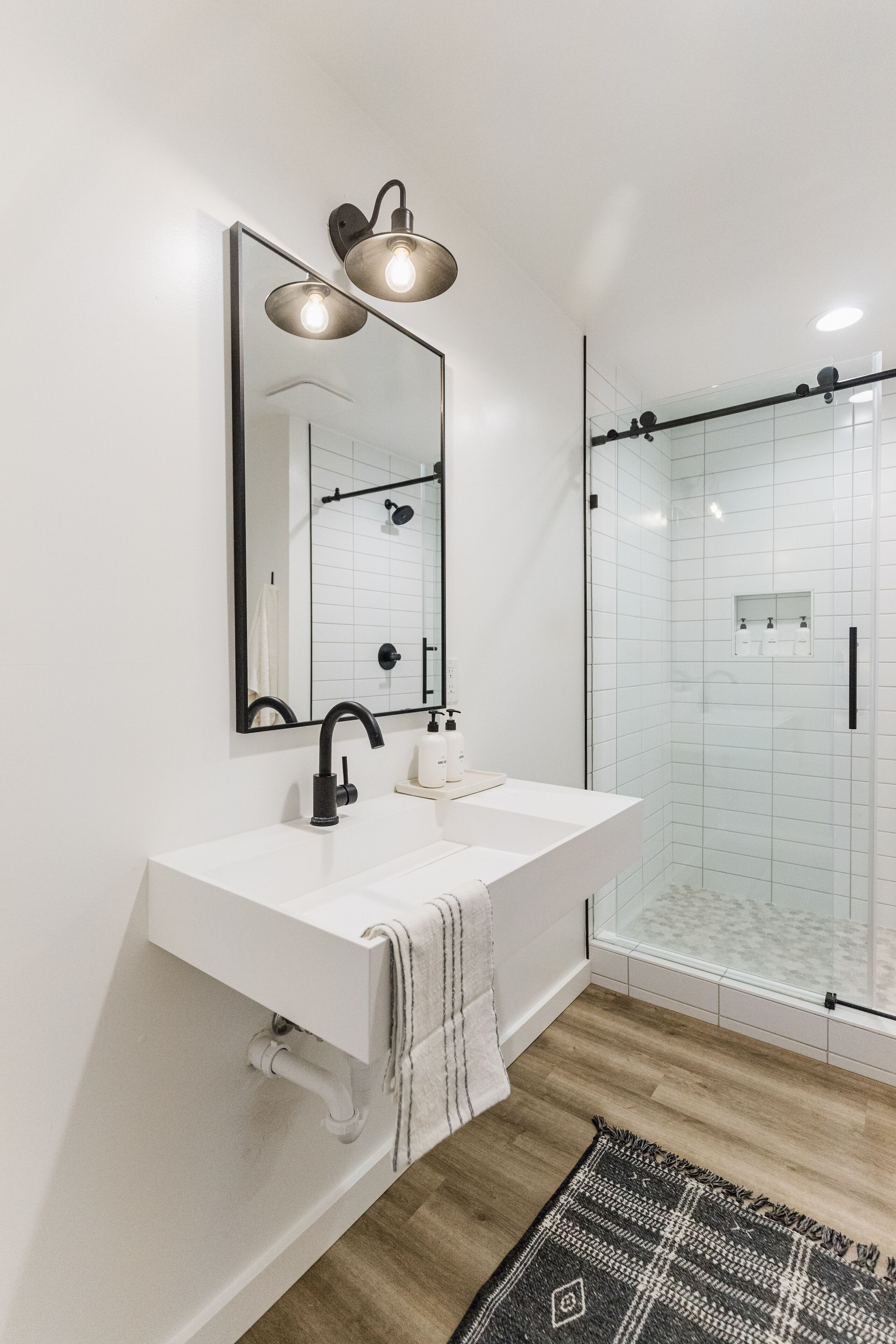 A modern bathroom with a white sink, black faucet, and a large mirror with two lights. There is a glass shower enclosure and a patterned rug on the floor.