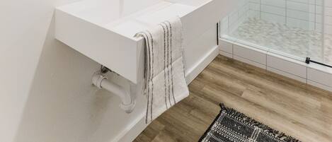 Un baño moderno con un lavamanos blanco, grifo negro y un espejo grande con dos luces. Cuenta con una cabina de ducha de vidrio y una alfombra estampada en el suelo.
