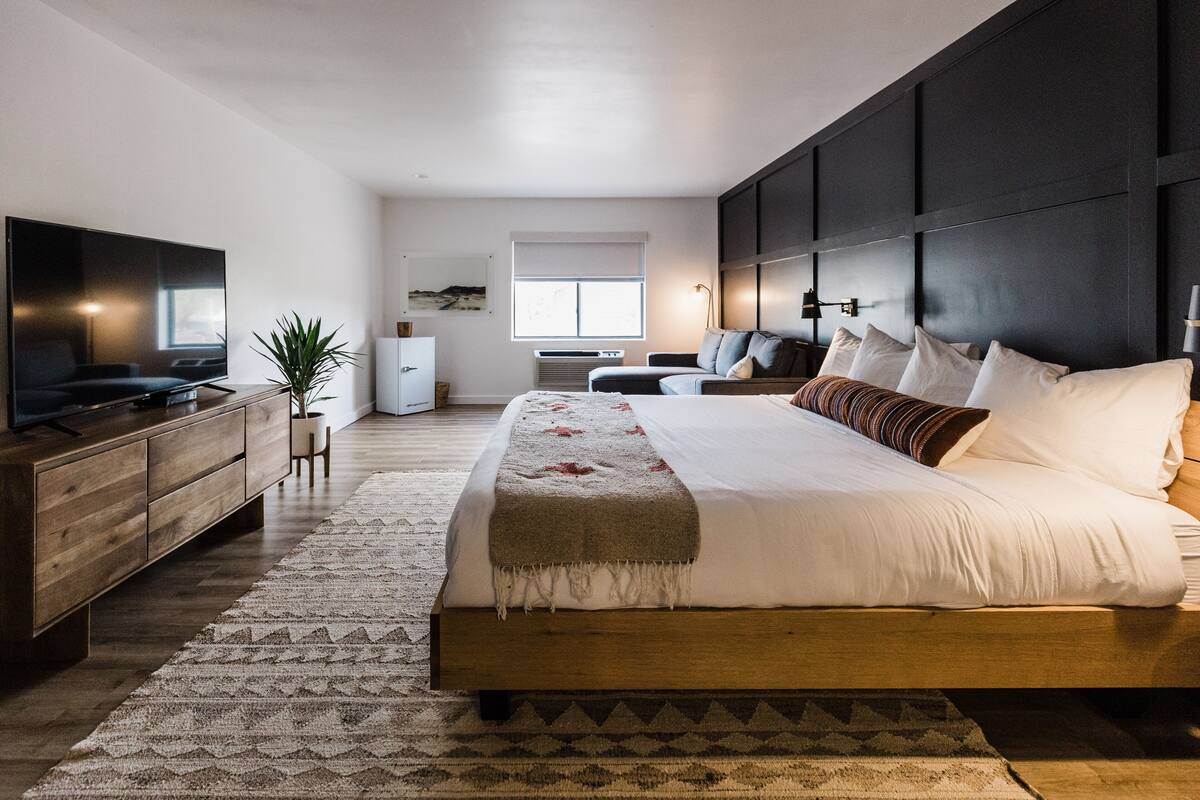 A modern bedroom with a large bed, a TV, a desk, and a plant.
