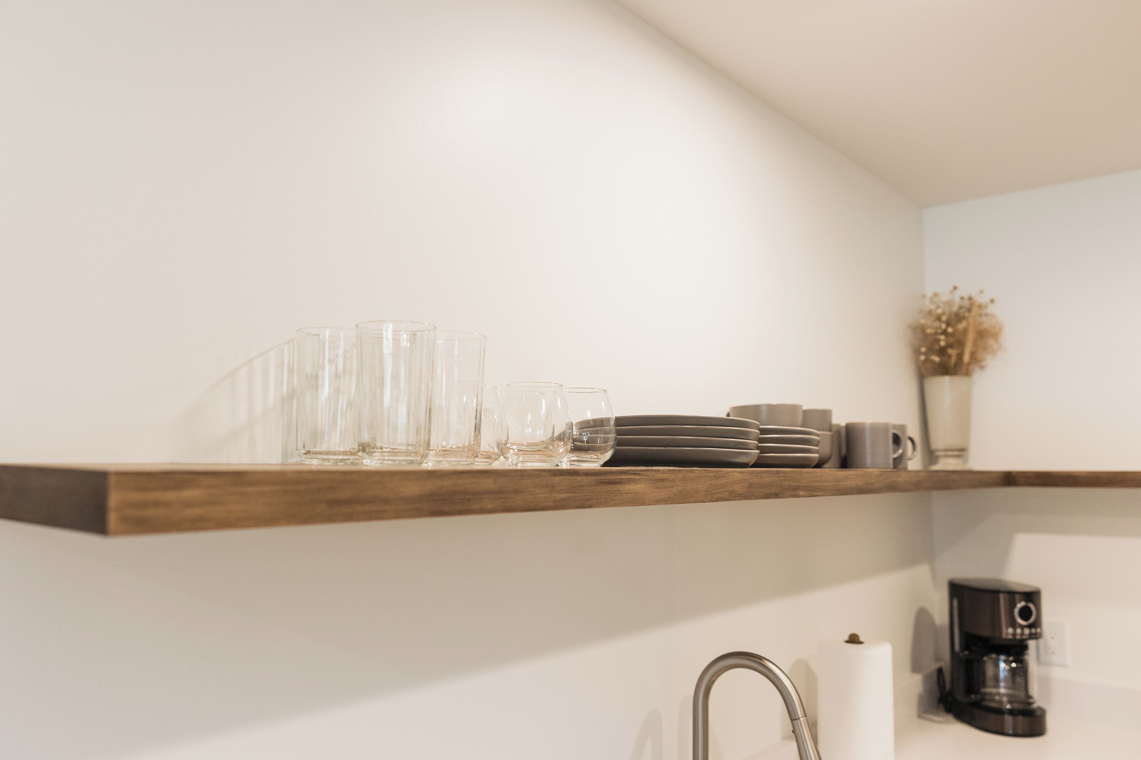 Una cocina con un estante de madera que contiene cristalería y platos, una cafetera y un jarrón con flores secas.
