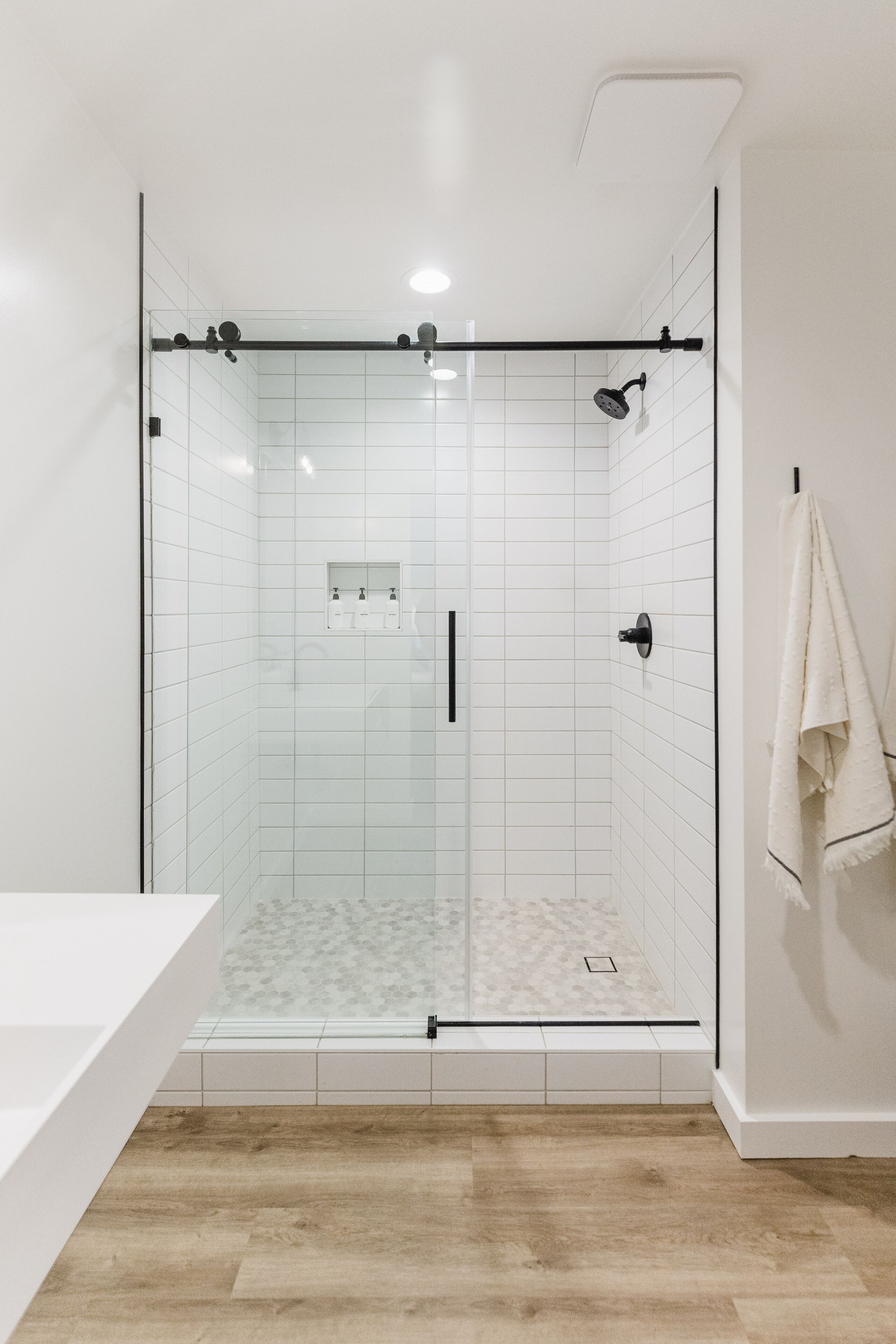 Un baño moderno con ducha de vidrio, paredes cubiertas de azulejos blancos, suelo de madera y encimera blanca.