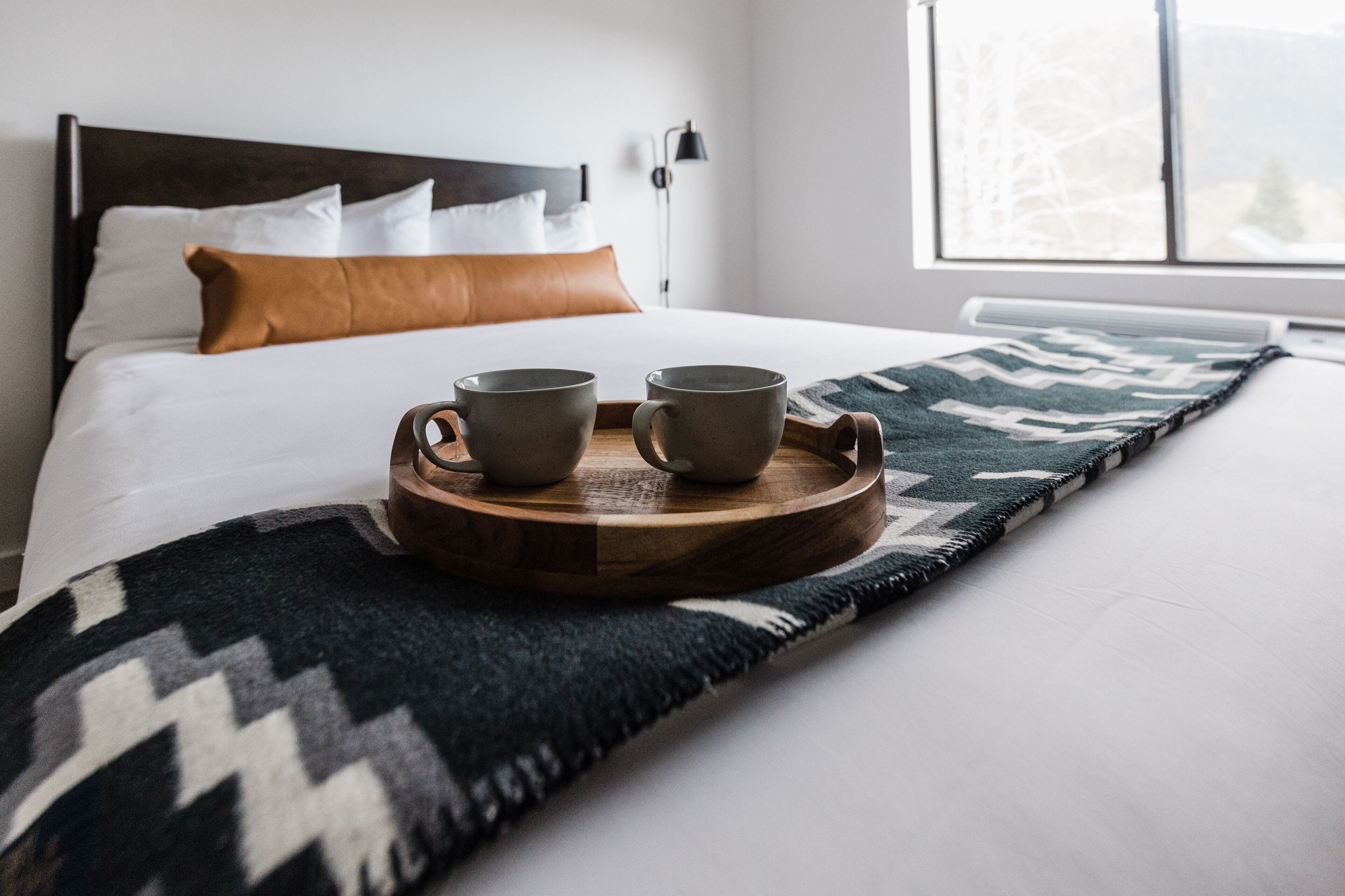 A neatly made bed with a wooden tray holding two mugs, a patterned throw blanket, and a view of a snowy landscape through a window.