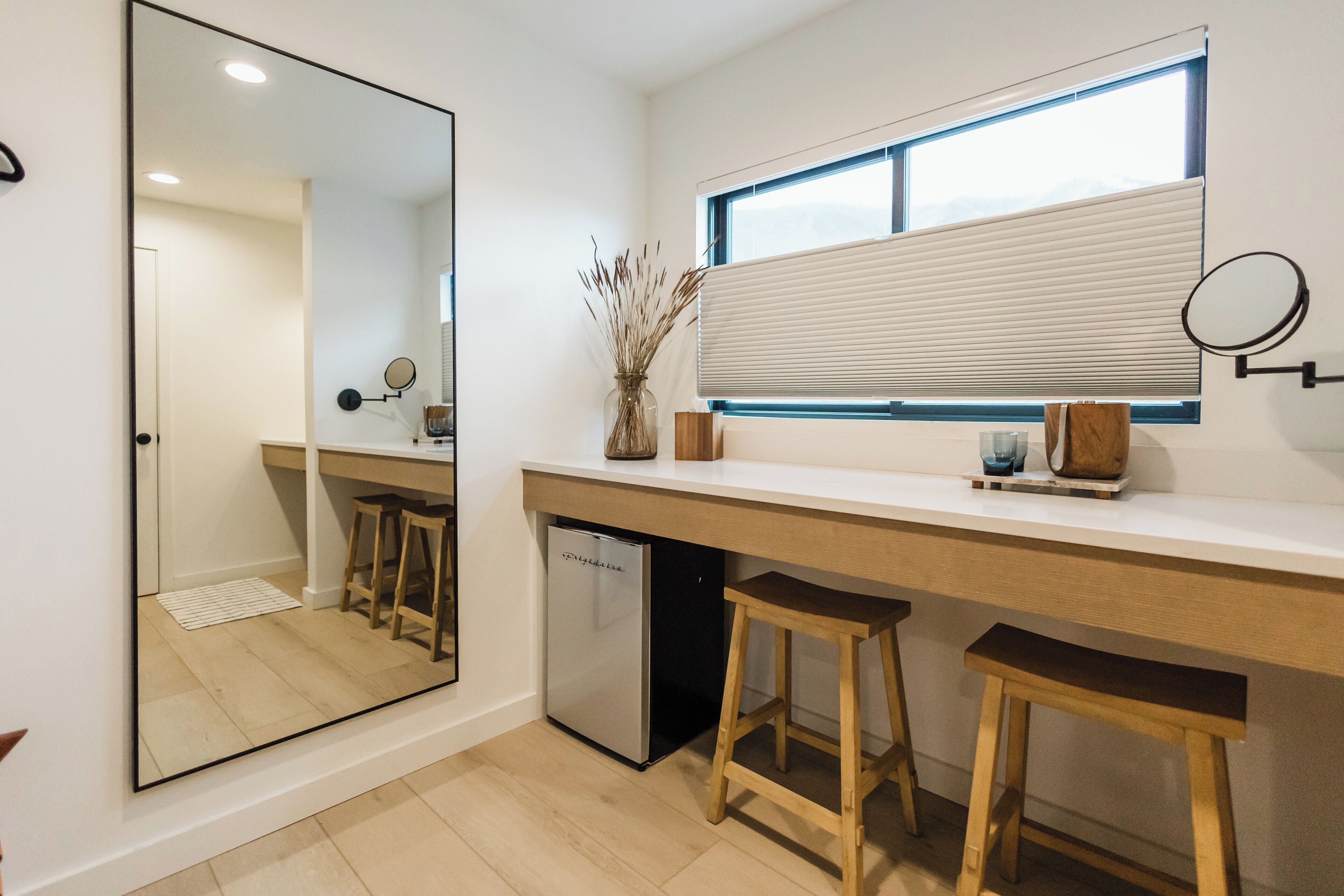 A modern kitchen with a large mirror, wooden stools, a built-in refrigerator, and a window with blinds.