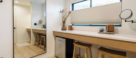 A modern kitchen with a large mirror, wooden stools, a built-in refrigerator, and a window with blinds.