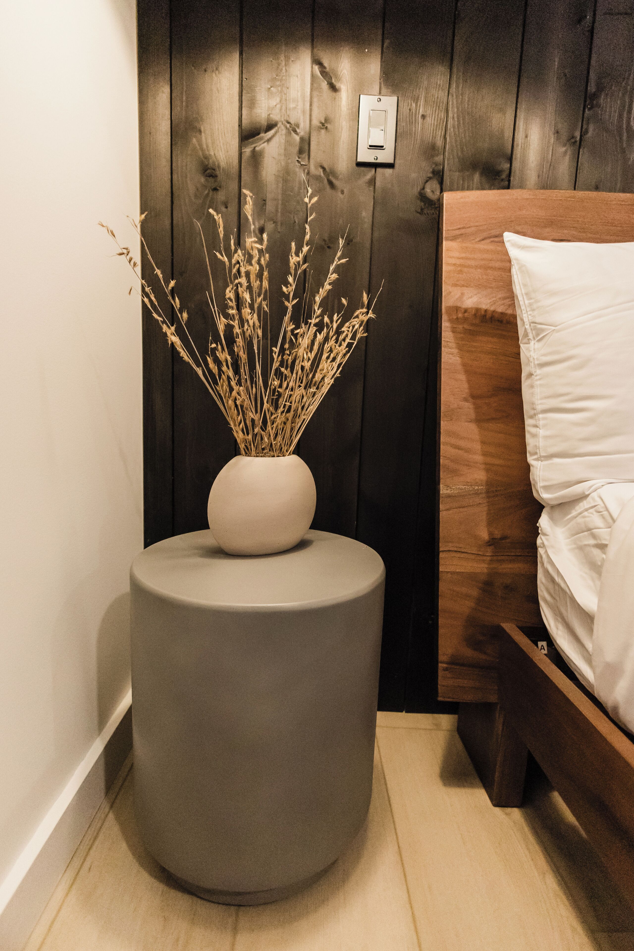 A corner of a room with a round grey ottoman, a white vase with dried plants, a wooden bed frame, and a dark wooden wall.