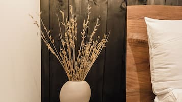 Rincón de una habitación con un otoman gris redondo, un jarrón blanco con plantas secas, un cabecero de cama de madera y una pared de madera oscura.