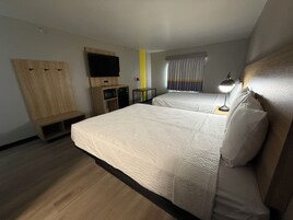 A hotel room with two beds, a TV mounted on the wall, a desk with a lamp, and a window with curtains.