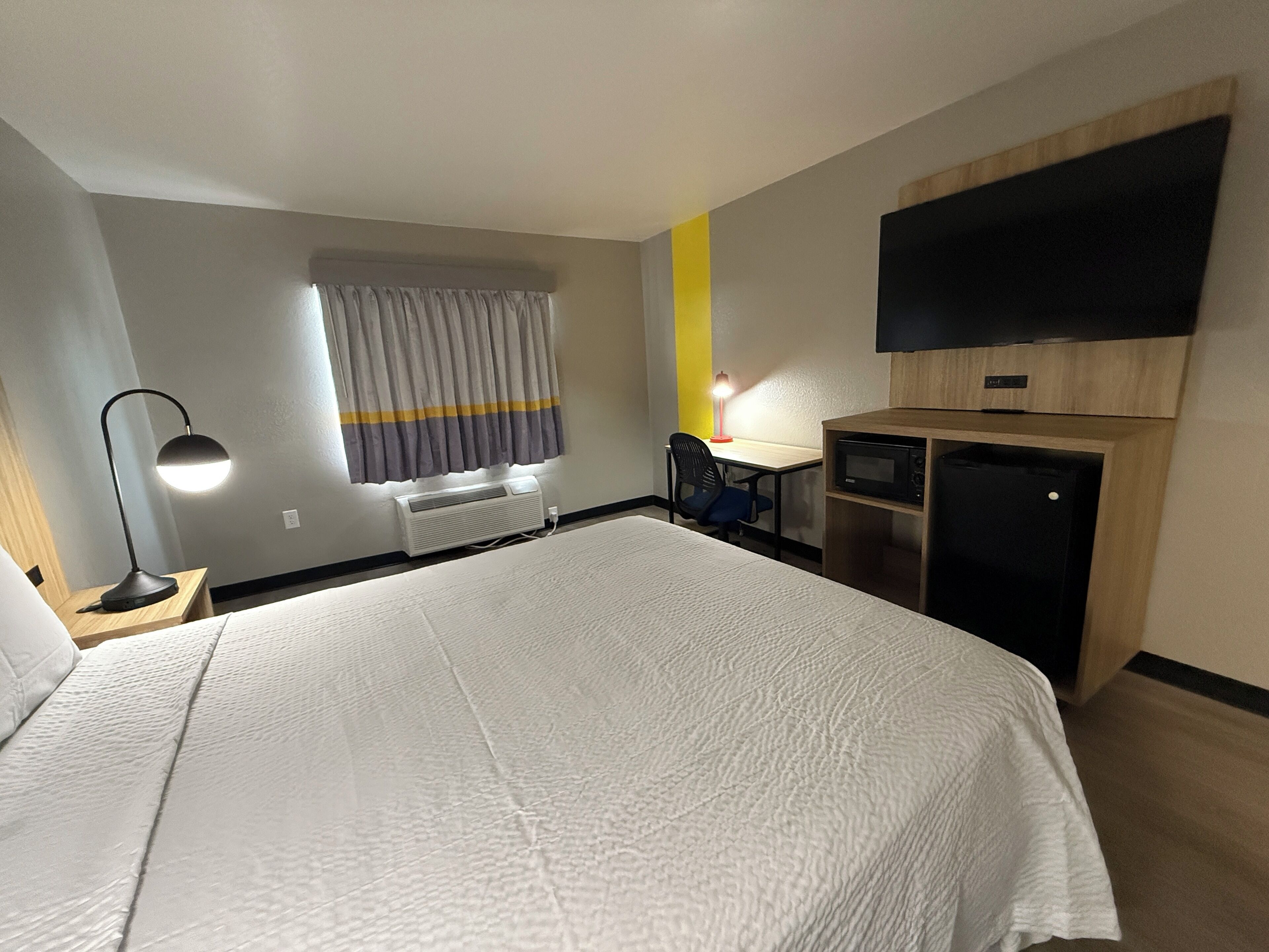 A hotel room with a large bed, a desk with a chair, a flat-screen TV mounted on the wall, and a window with curtains.
