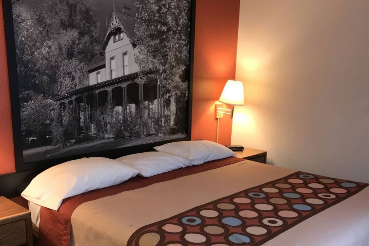 Une chambre d’hôtel avec un grand lit, une table de chevet, une lampe fixée au mur et un tableau encadré au-dessus du lit.