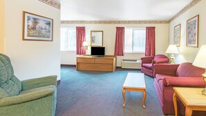 Una habitación de hotel con un sillón verde, un sofá rojo, una mesa de centro de madera, un televisor sobre un mueble de madera y dos ventanas con cortinas rojas.
