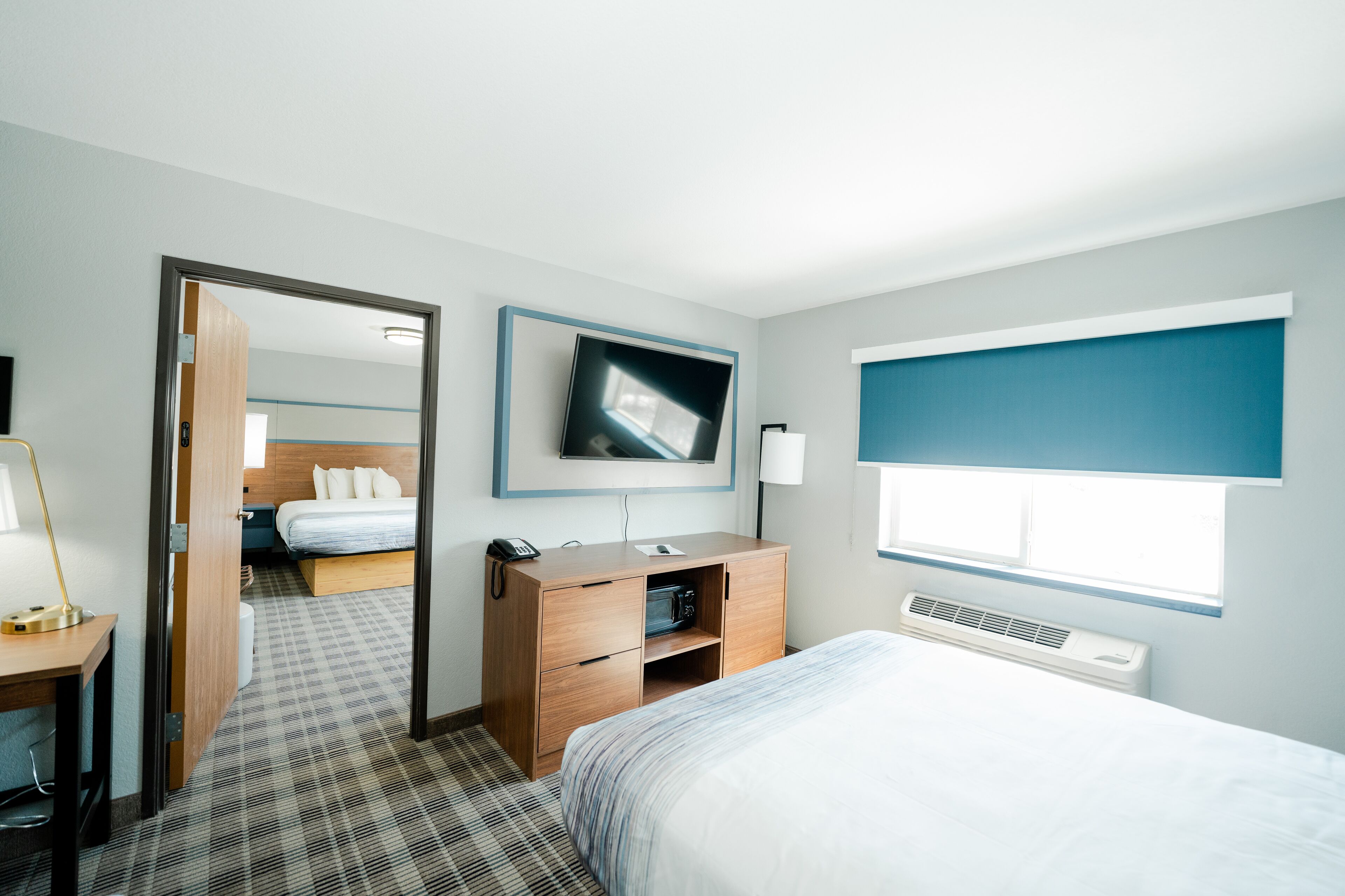 A hotel room with a bed, a desk with a lamp, a TV mounted on the wall, and a window with blinds.