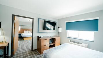 A hotel room with a bed, a desk with a lamp, a TV mounted on the wall, and a window with blinds.
