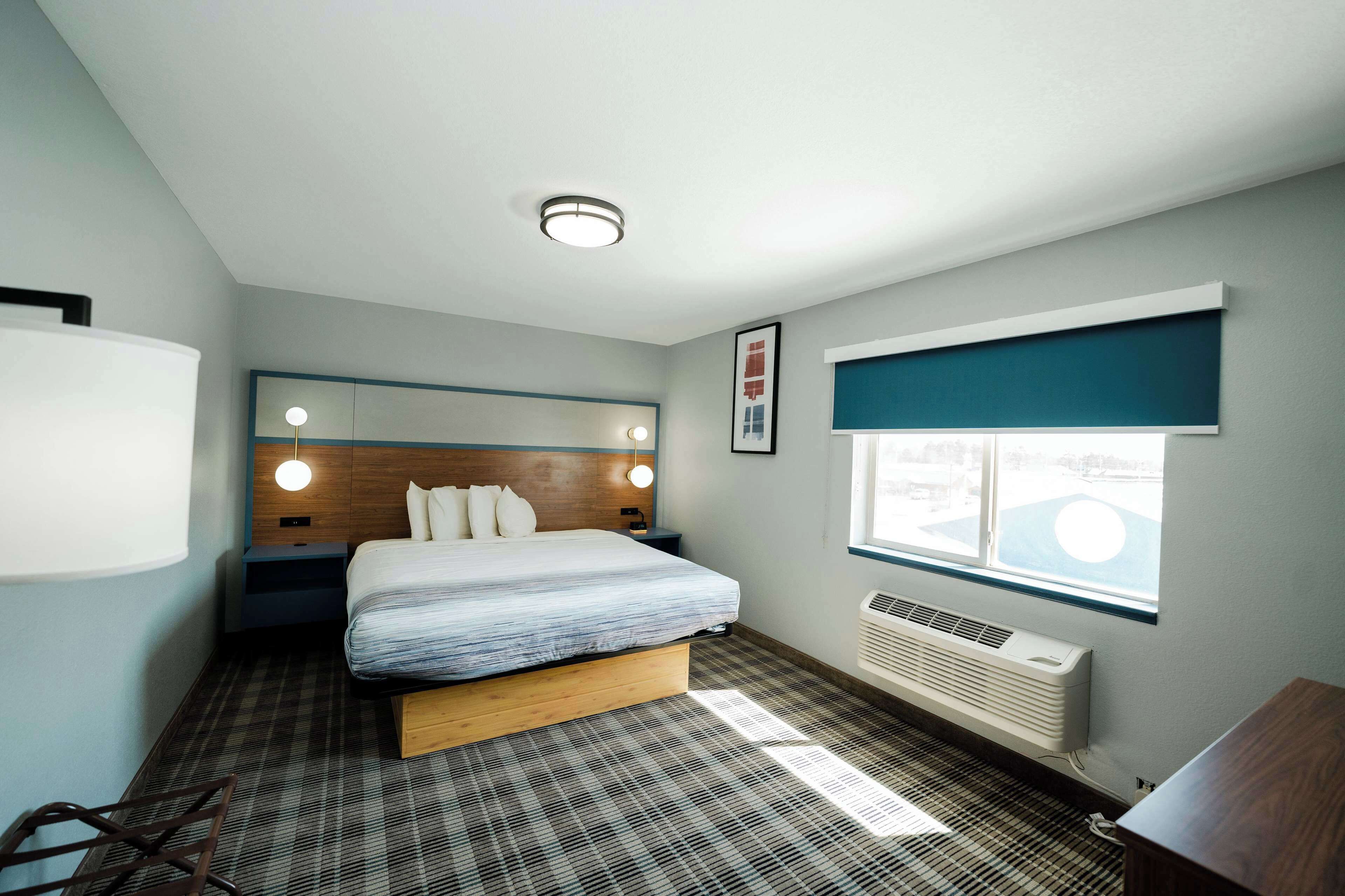 Une chambre d’hôtel moderne équipée d’un lit, de tables de chevet, d’une fenêtre avec stores, d’un radiateur fixé au mur et d’un luminaire au plafond.