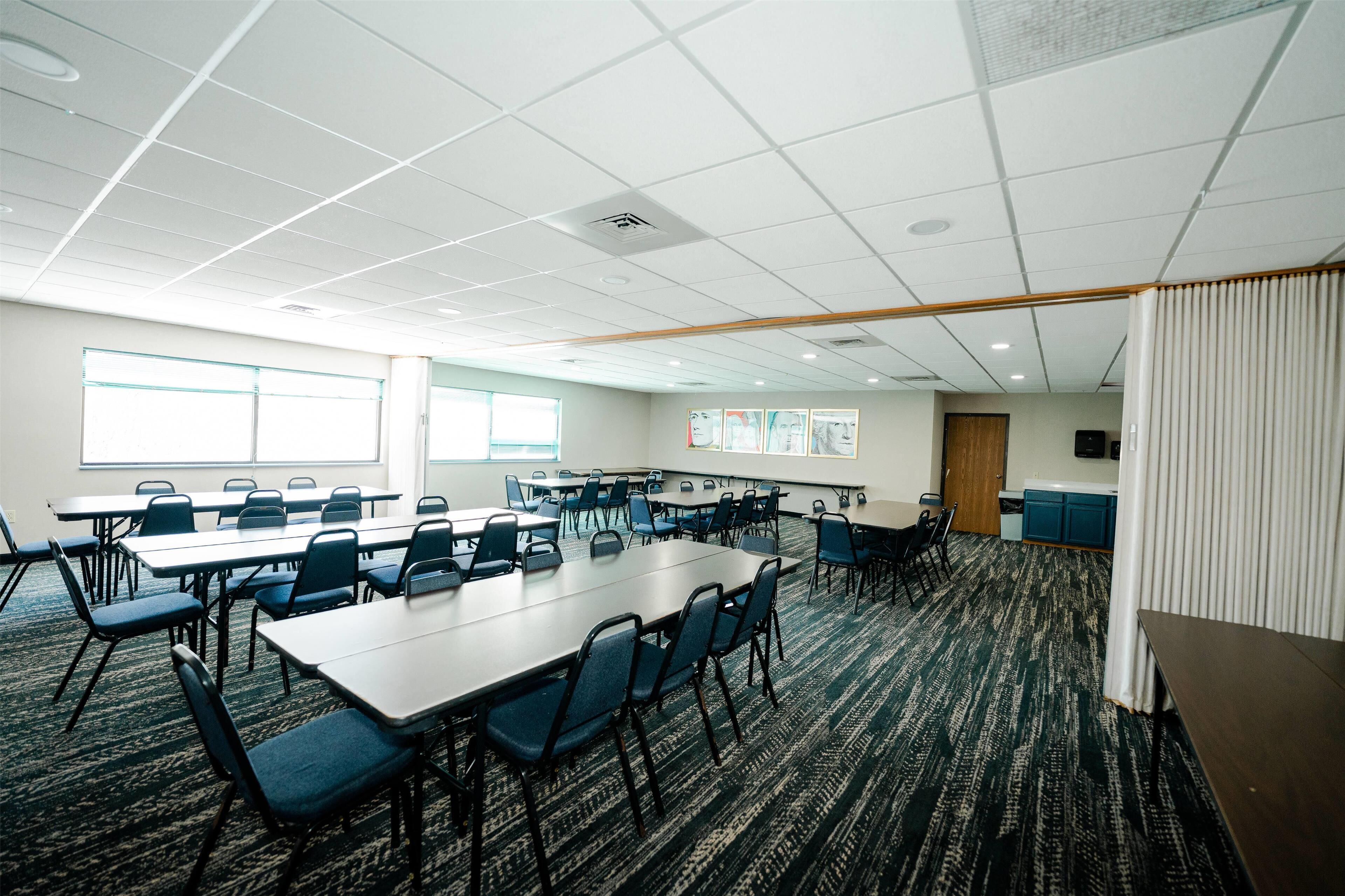 A conference room with multiple tables and chairs, a kitchenette, and a large window.