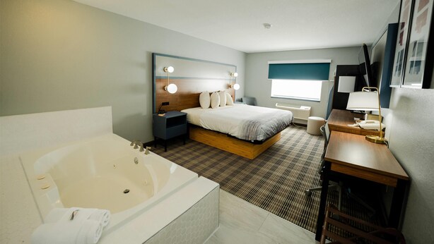 Habitación de hotel moderna con una cama grande, un área de estar independiente con un escritorio y una silla, un baño separado con bañera y un ventanal que proporciona luz natural.