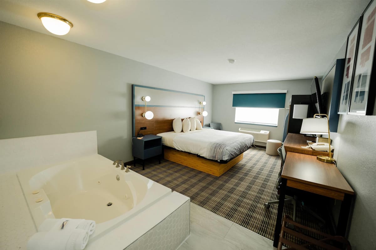 A modern hotel room with a large bed, a separate sitting area with a desk and chair, a separate bathroom with a bathtub, and a large window providing natural light.