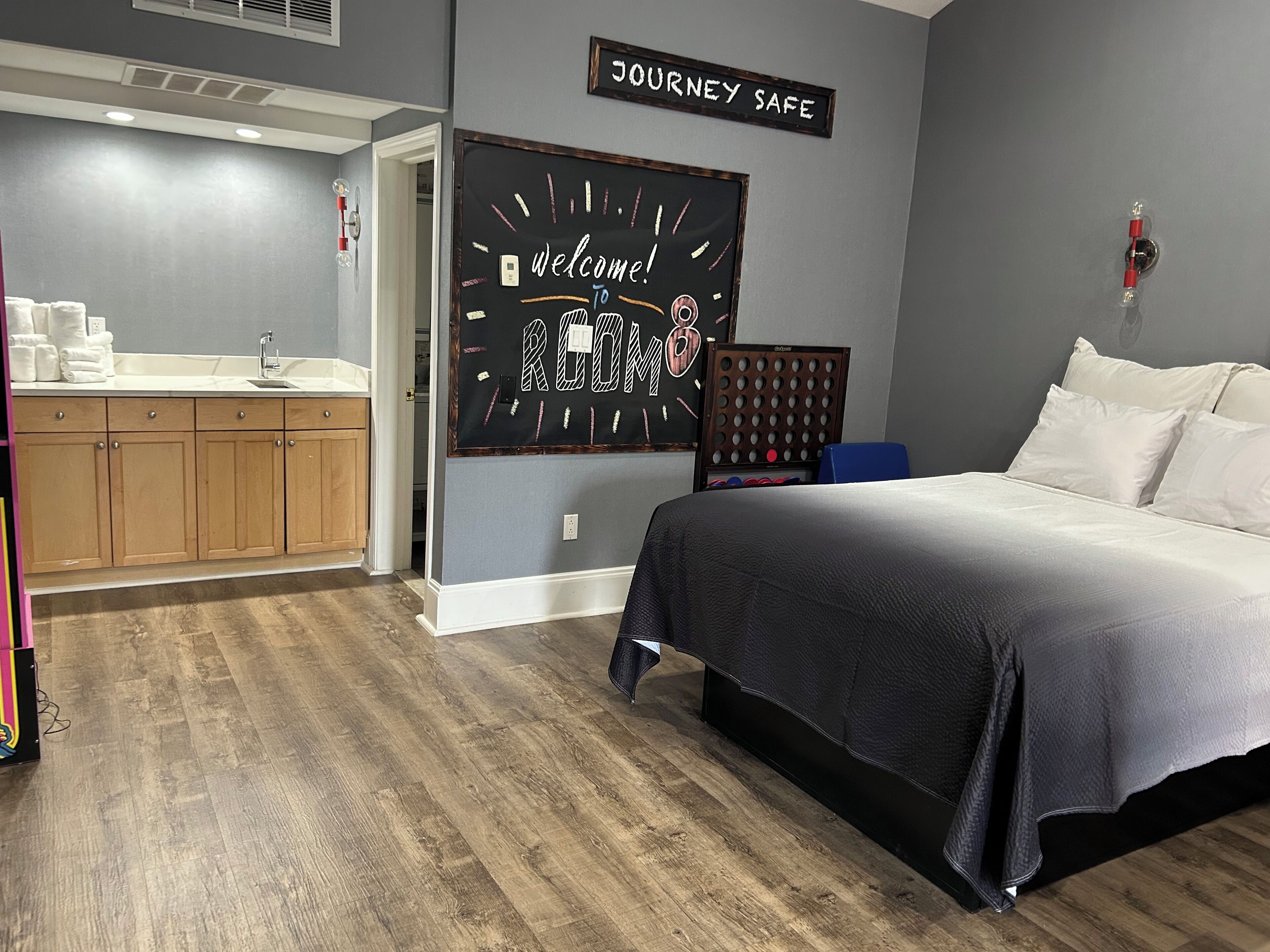 A hotel room with a bed, a small desk, a kitchenette, and a sign that reads "WELCOME TO ROOM 8".