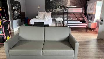 Une chambre moderne avec un canapé gris, des lits superposés, une affiche ancienne représentant un village et un panneau indiquant « Bon voyage ».