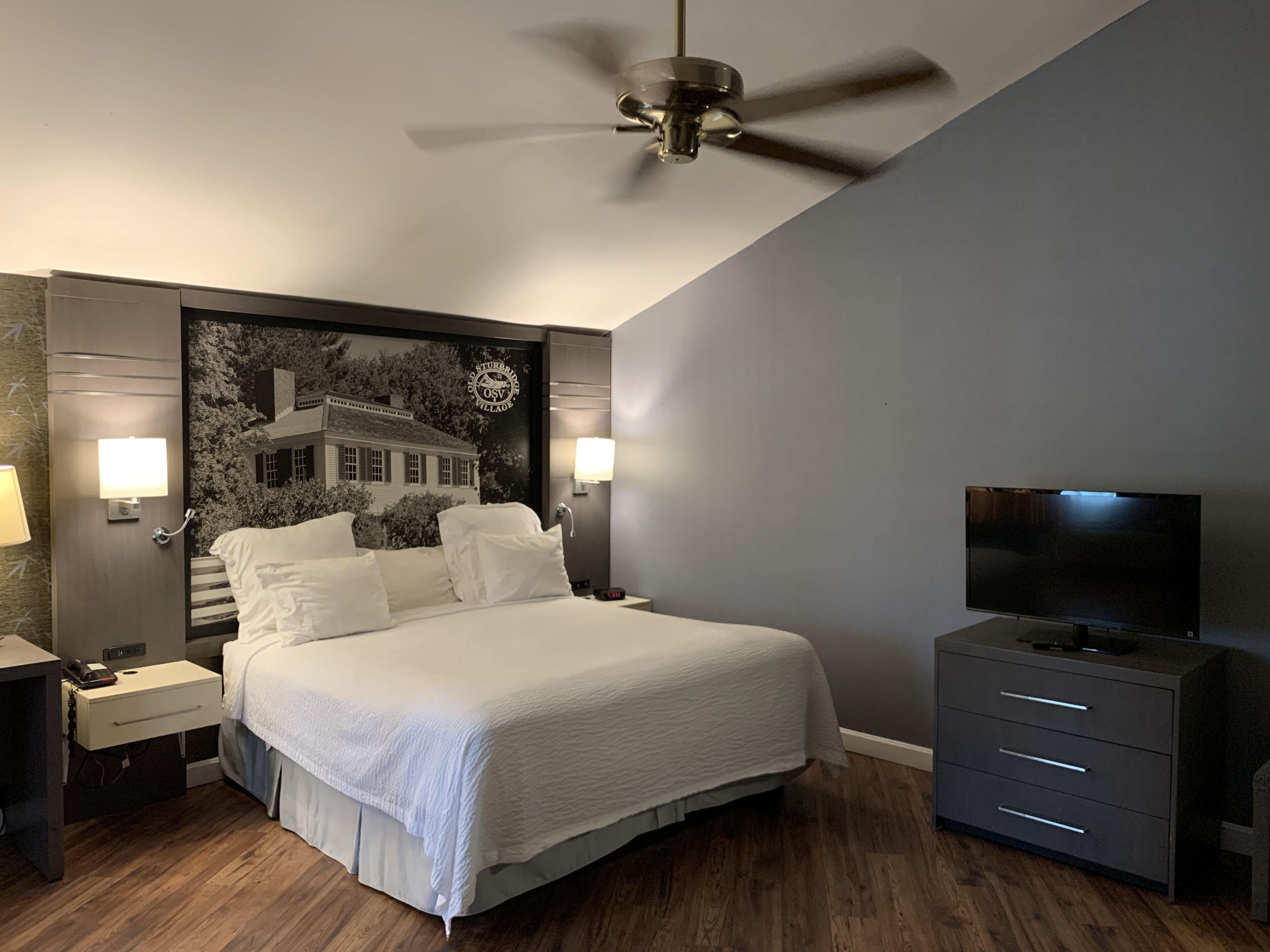 A modern bedroom with a large bed, a ceiling fan, a TV, and a nightstand with a lamp.