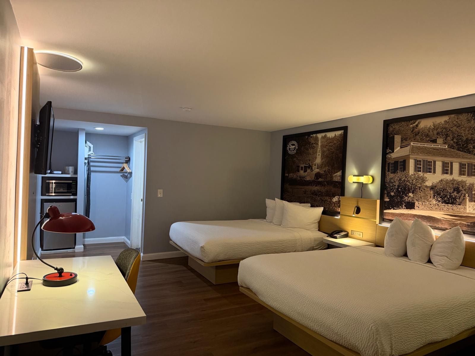 A hotel room with a bed, a desk, a chair, a lamp, a nightstand, and a wall-mounted TV.