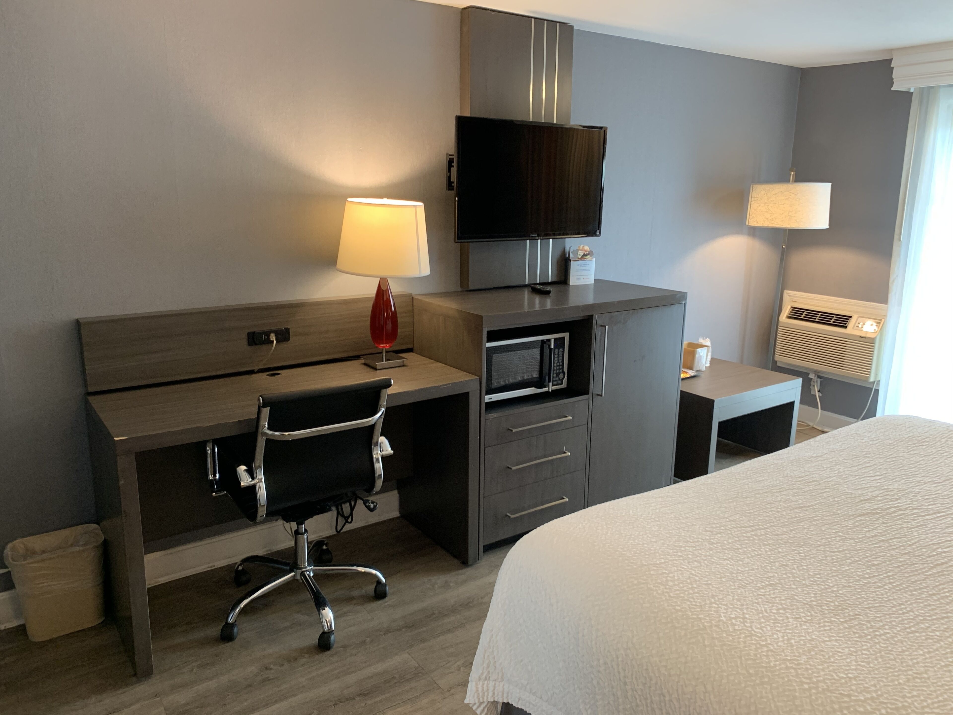 A hotel room with a bed, a desk with a chair, a TV, a microwave, and two lamps.