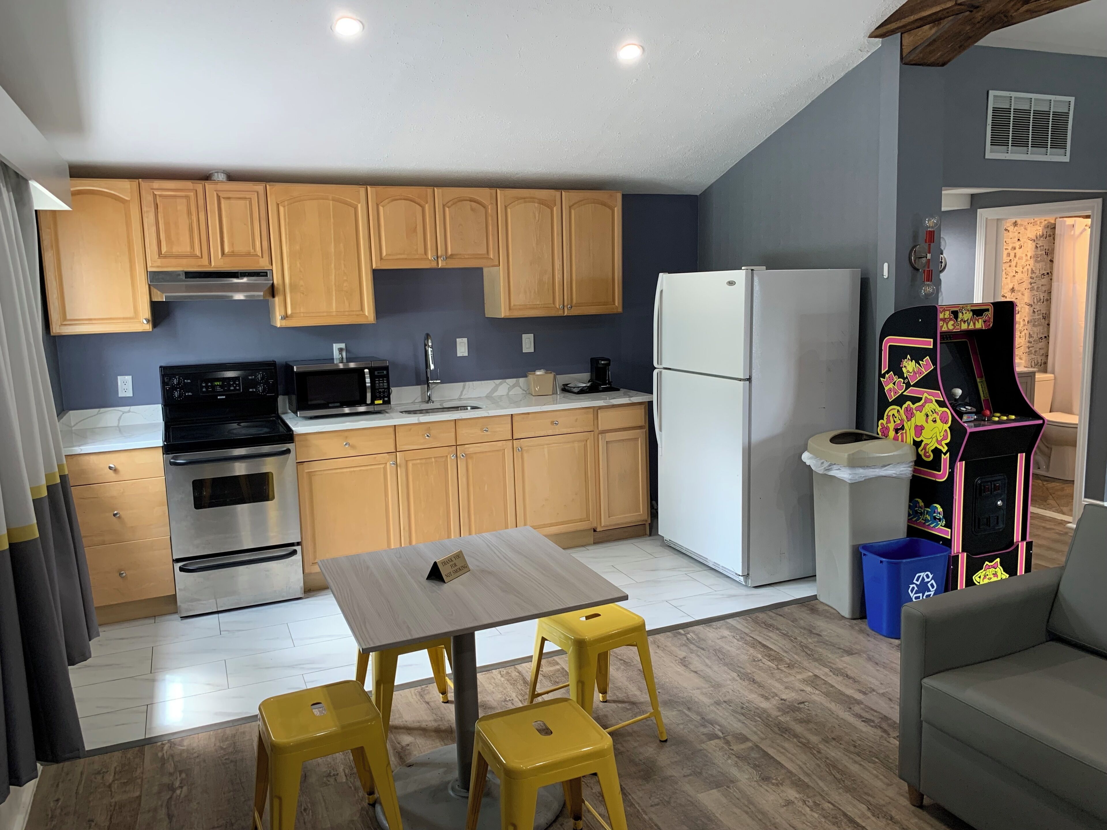 A compact kitchen with wooden cabinets, a microwave, stove, and a refrigerator. There is a small dining table with yellow stools and a grey sofa.