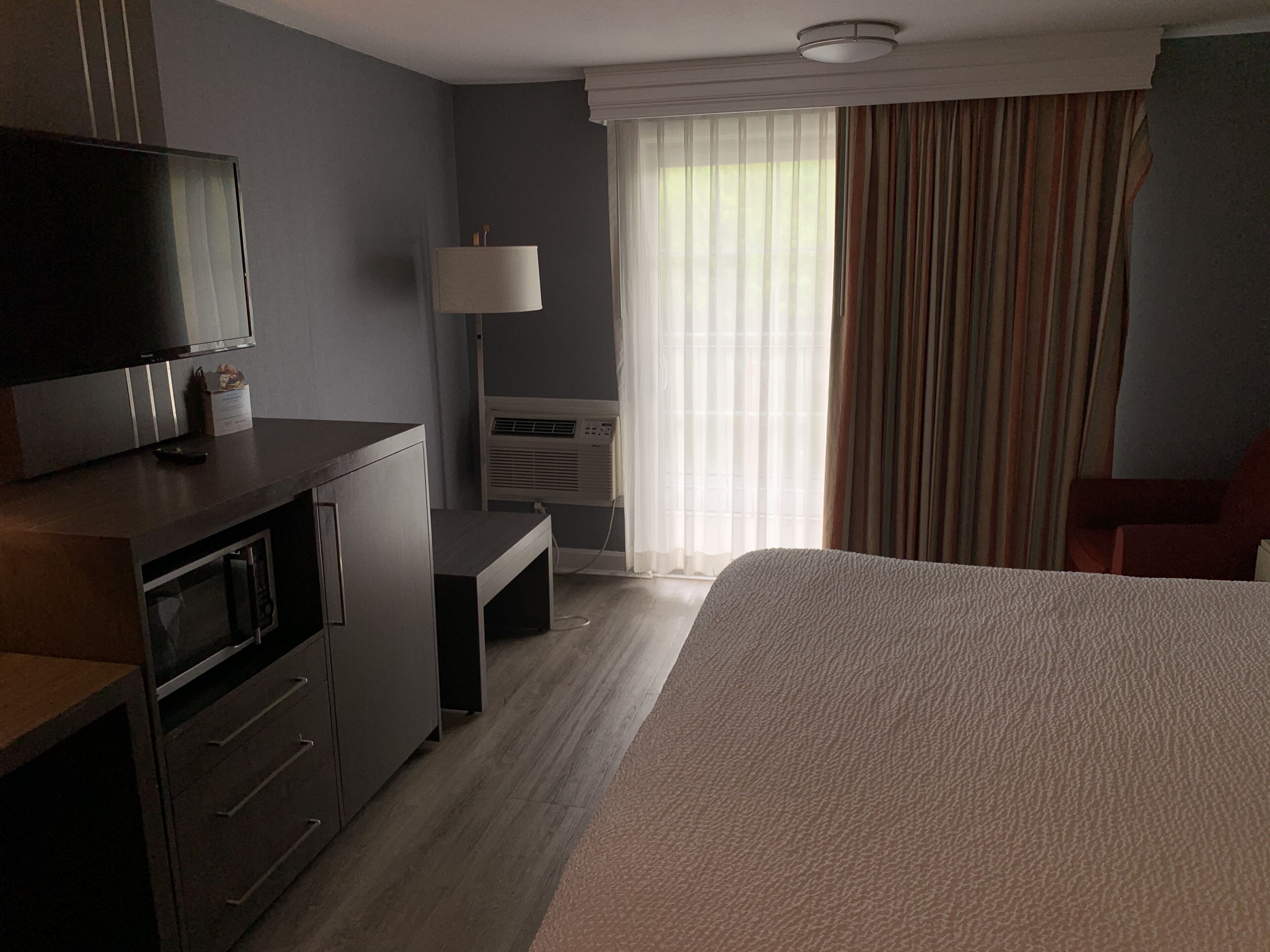Habitación de hotel con cama, escritorio, televisión, aire acondicionado y ventana con cortinas.