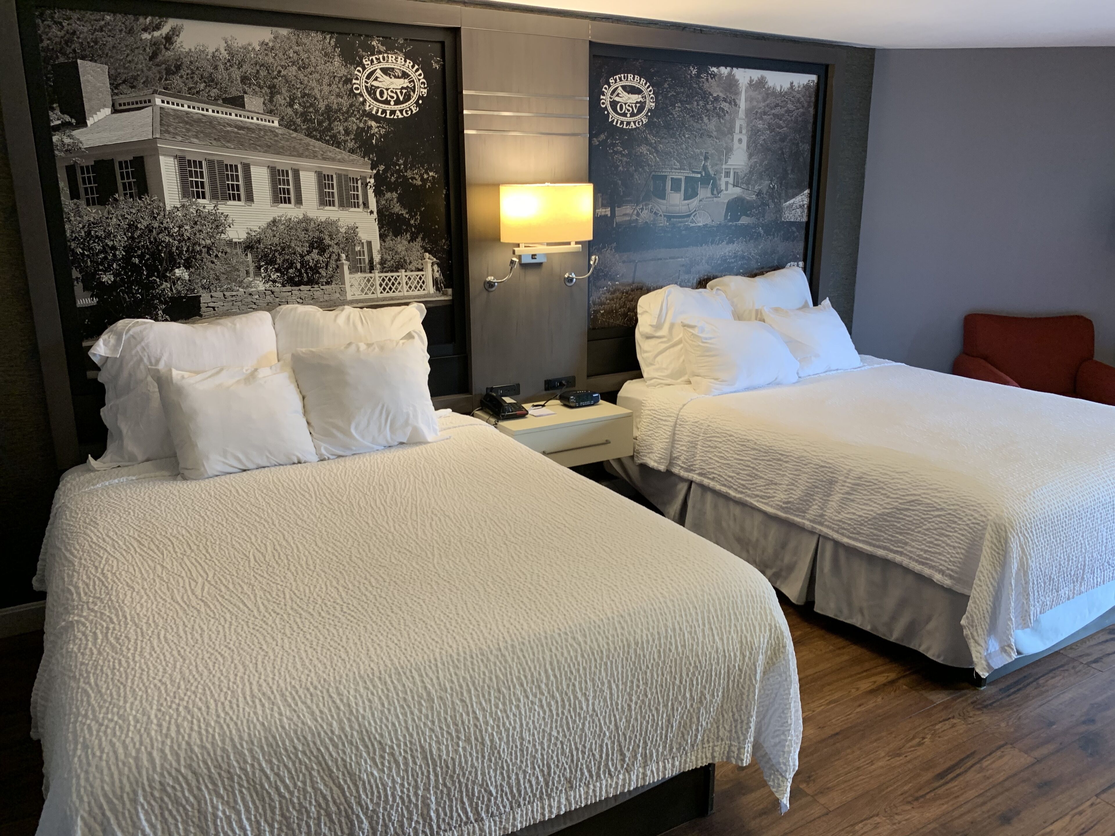 Habitación de hotel con dos camas, una mesita de noche, una lámpara y un cuadro grande con un edificio histórico.