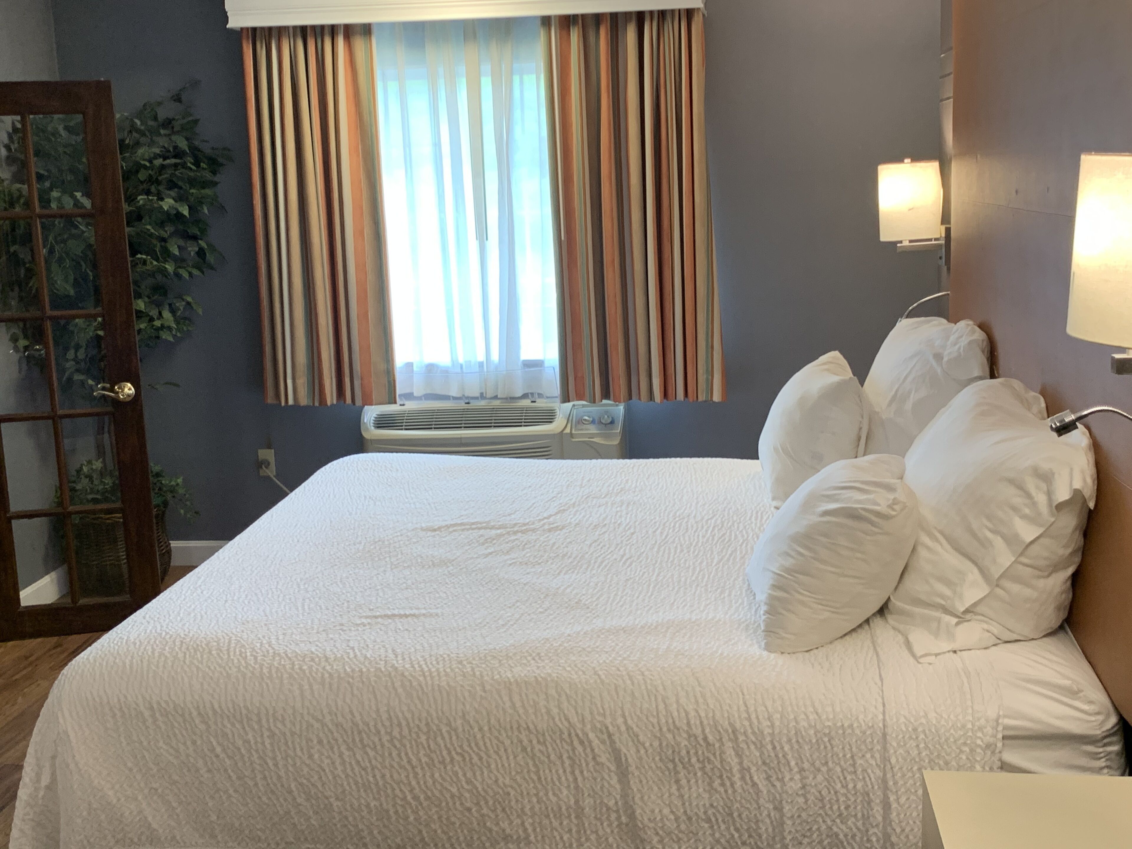 A hotel room with a large bed, a window with curtains, an air conditioning unit, and a bedside lamp.