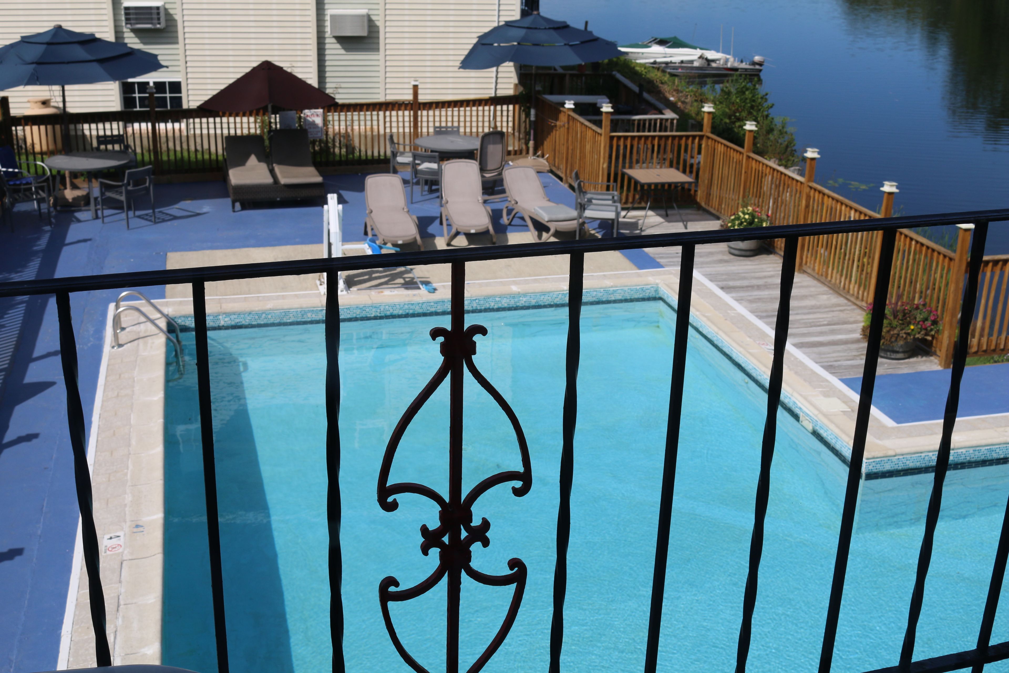 Une piscine entourée d’une clôture en métal décorative, de chaises longues et de parasols, avec une terrasse en bois et une vue sur une étendue d’eau.