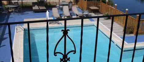 A pool with a decorative metal fence, surrounded by lounge chairs and umbrellas, with a wooden deck and a view of a body of water.