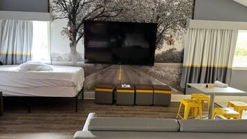 A modern hotel room with a large flat-screen TV mounted on a wall with a tree and road mural, a bed, a sofa, and a small dining area with yellow stools.