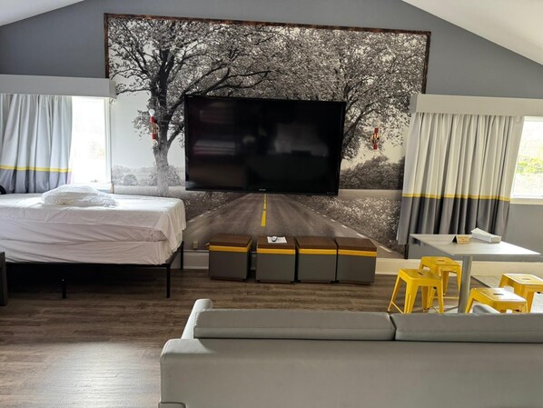 A modern hotel room with a large flat-screen TV mounted on a wall with a tree and road mural, a bed, a sofa, and a small dining area with yellow stools. - Super 8 by Wyndham Sturbridge (Sturbridge)