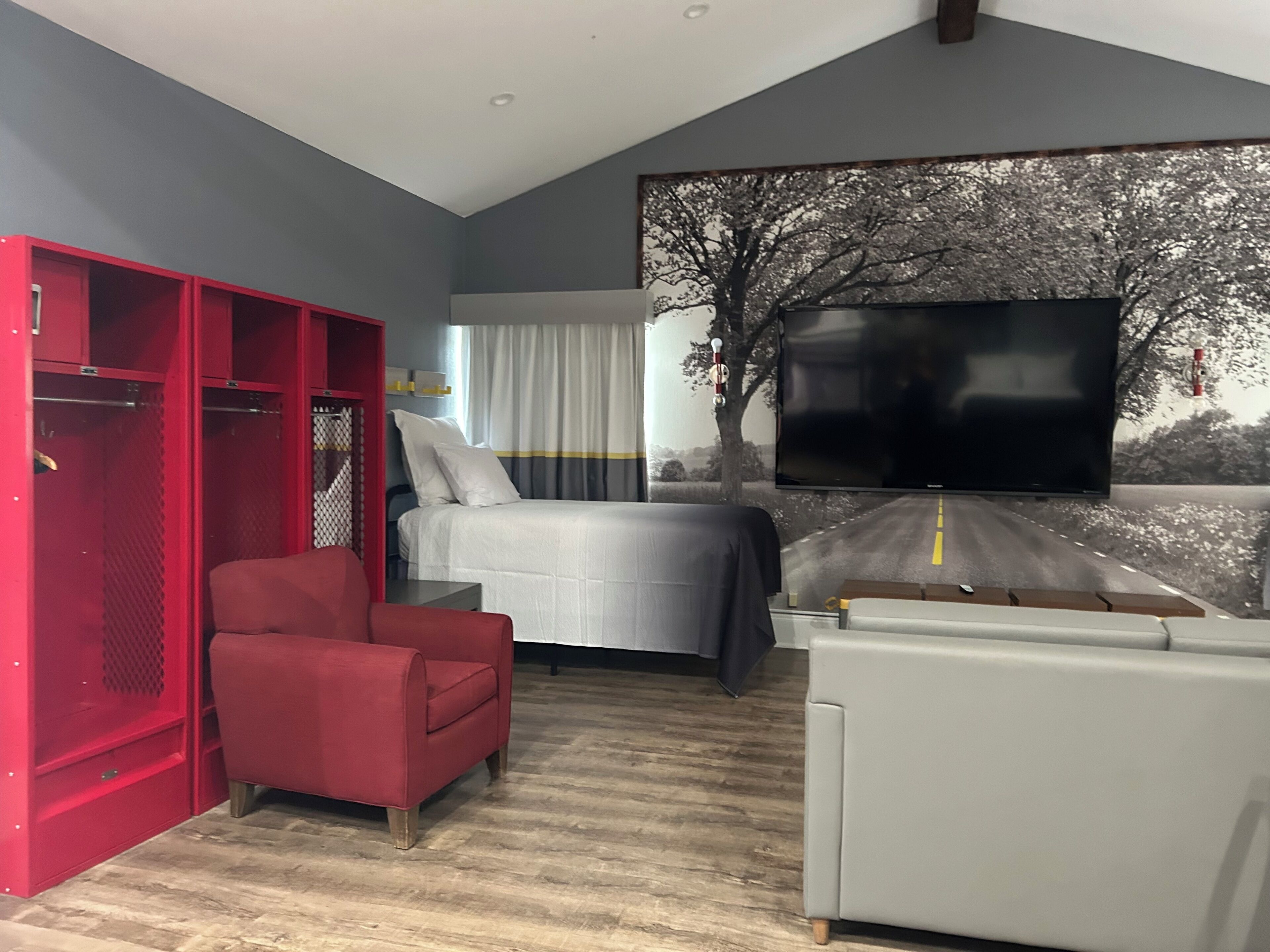 A modern hotel room with a bed, a red armchair, a grey sofa, a TV, and a large window with a road and tree mural.