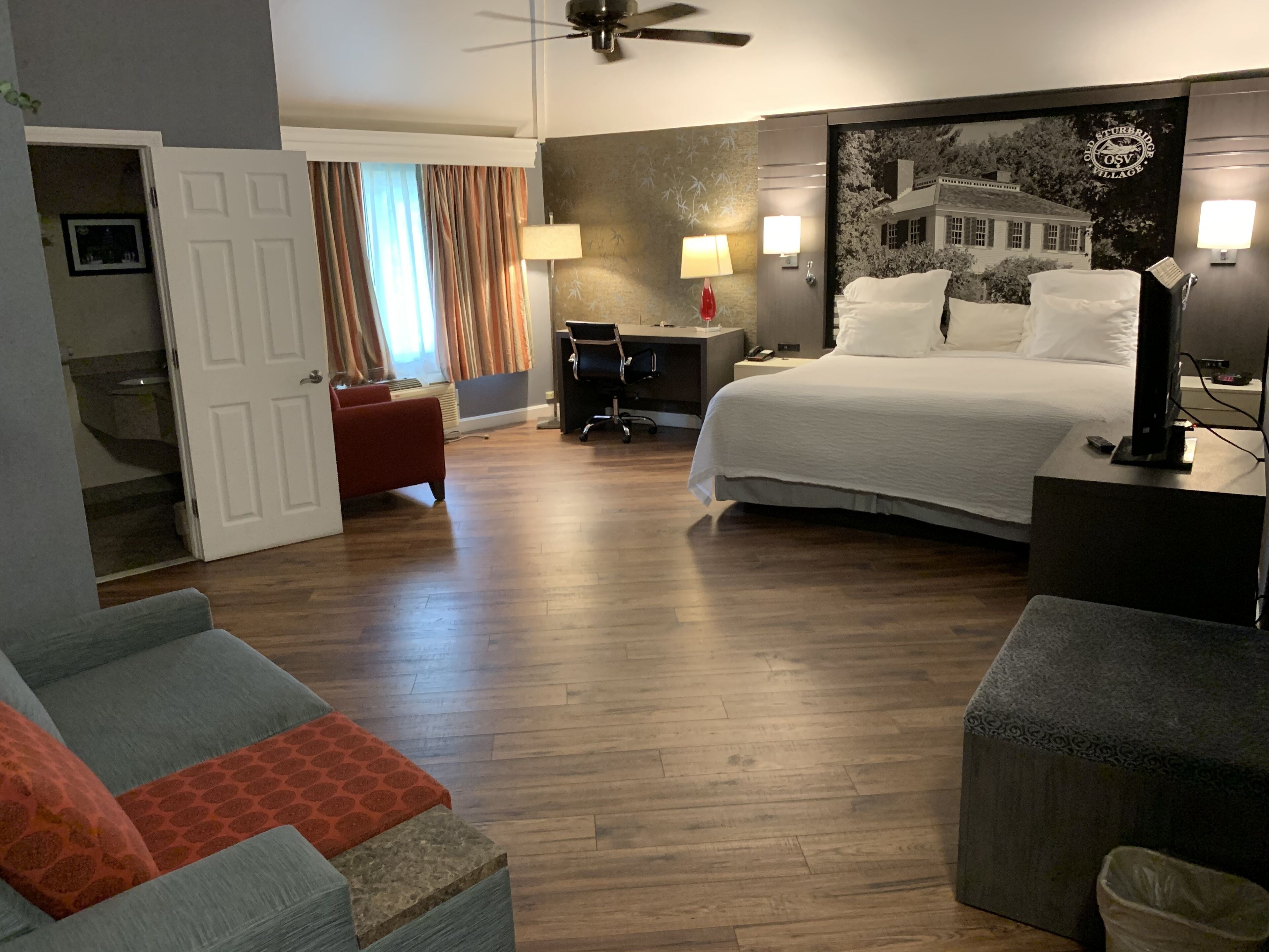 A hotel room with a bed, desk, chair, and a view of a house through a window.
