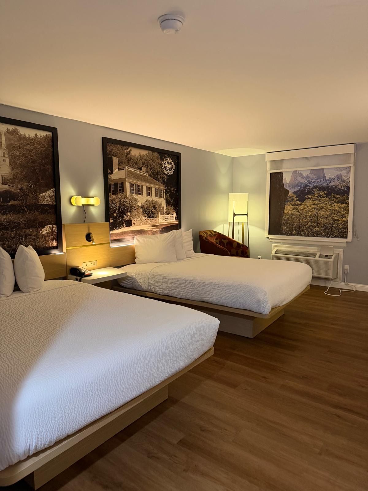A hotel room with two beds, a desk, a chair, a lamp, and a large window with a mountain view.