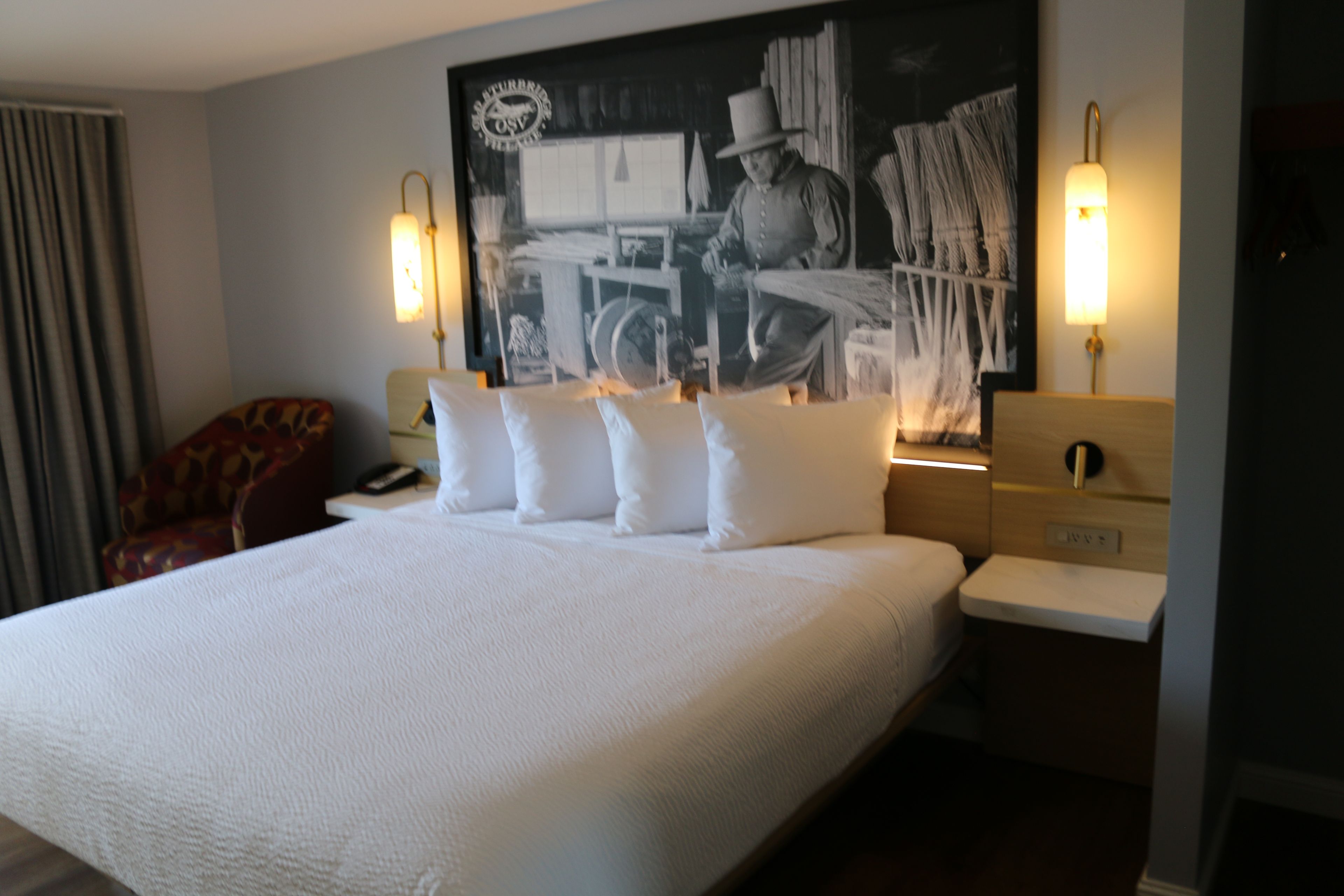 Una habitación de hotel con una cama grande, un cabecero con un cuadro, dos lámparas de noche y una silla con un cojín estampado.