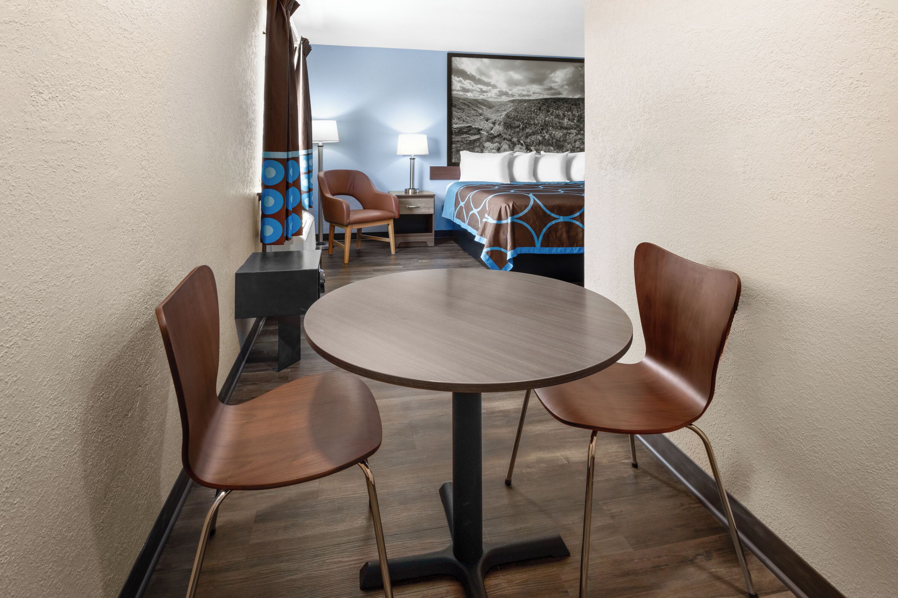 A compact hotel room with a round dining table and two chairs, a bed with a blue and brown bedspread, a nightstand with a lamp, and a wall-mounted TV.