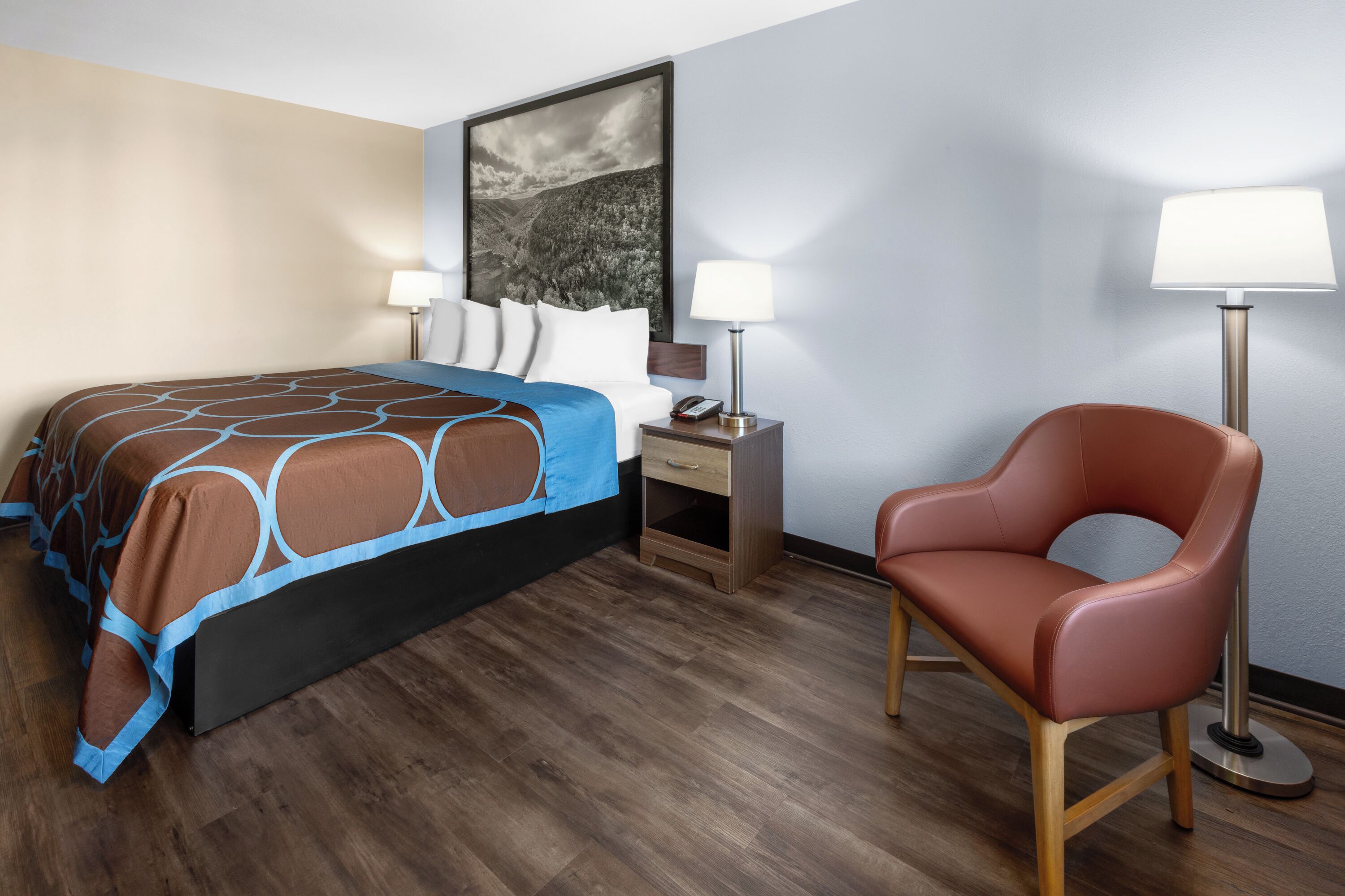 Una habitació d'hotel amb un llit, una taula de nit, una làmpada, una cadira i un quadre a la paret.