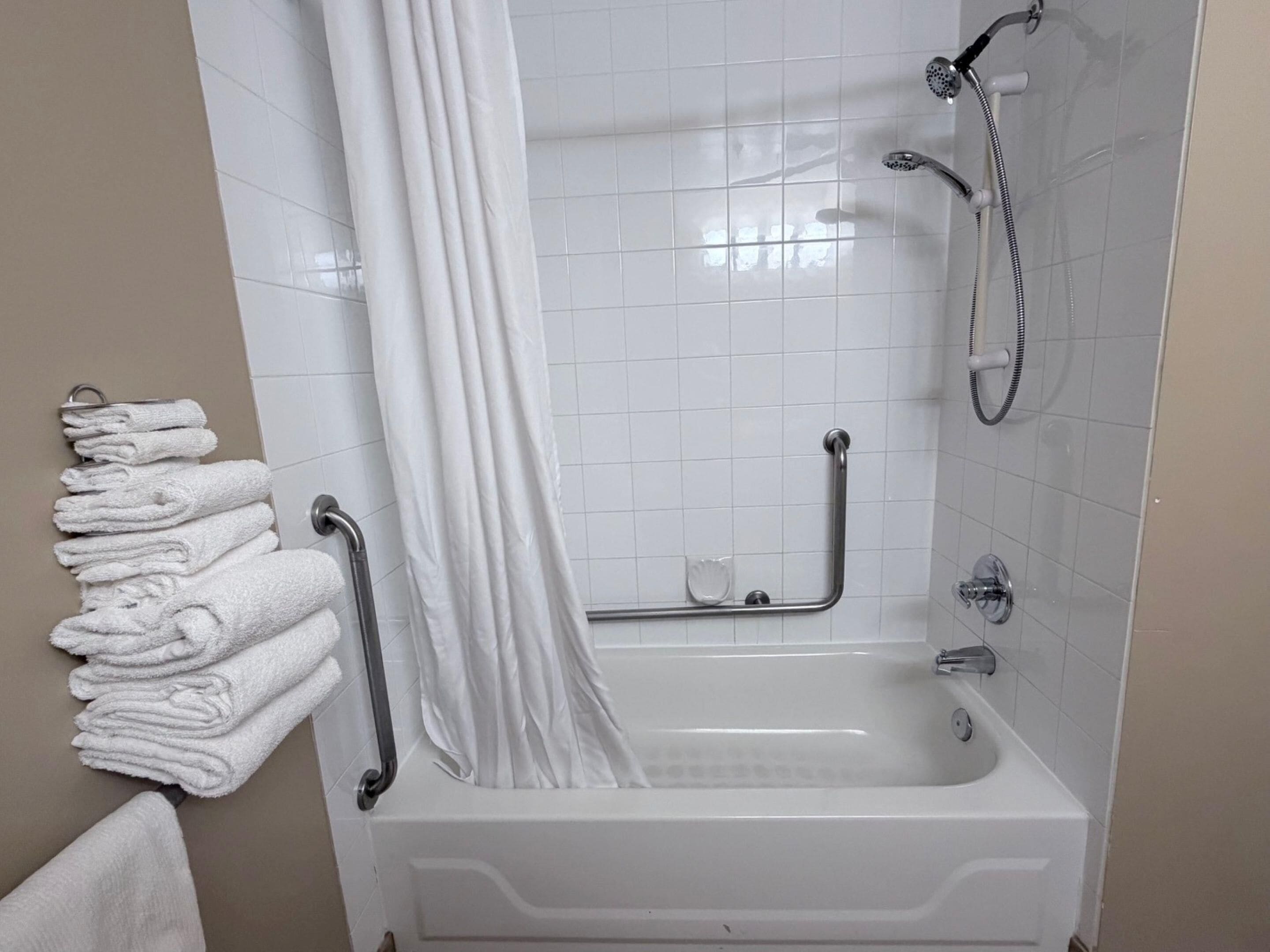 A bathroom with a bathtub, a shower, a handrail, and a towel rack with folded towels.
