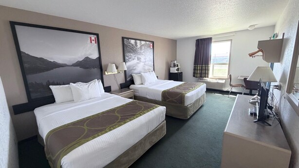 A hotel room with two beds, a desk, a chair, a window with curtains, a mounted TV, and a wall-mounted lamp.