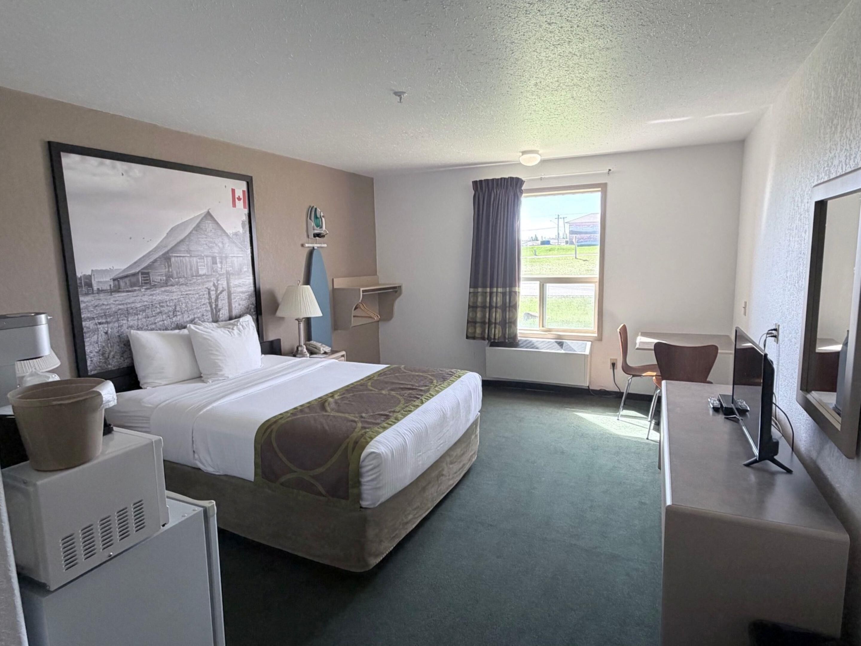 Une chambre d’hôtel comprenant un lit, un bureau, une chaise, une fenêtre avec des rideaux, une lampe, un radiateur et un tableau au mur.