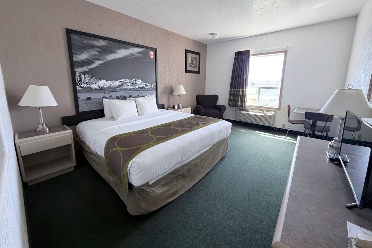 Une chambre d’hôtel avec un grand lit, deux tables de chevet avec des lampes, un bureau avec une chaise, une fenêtre avec des rideaux et un tableau accroché au mur.
