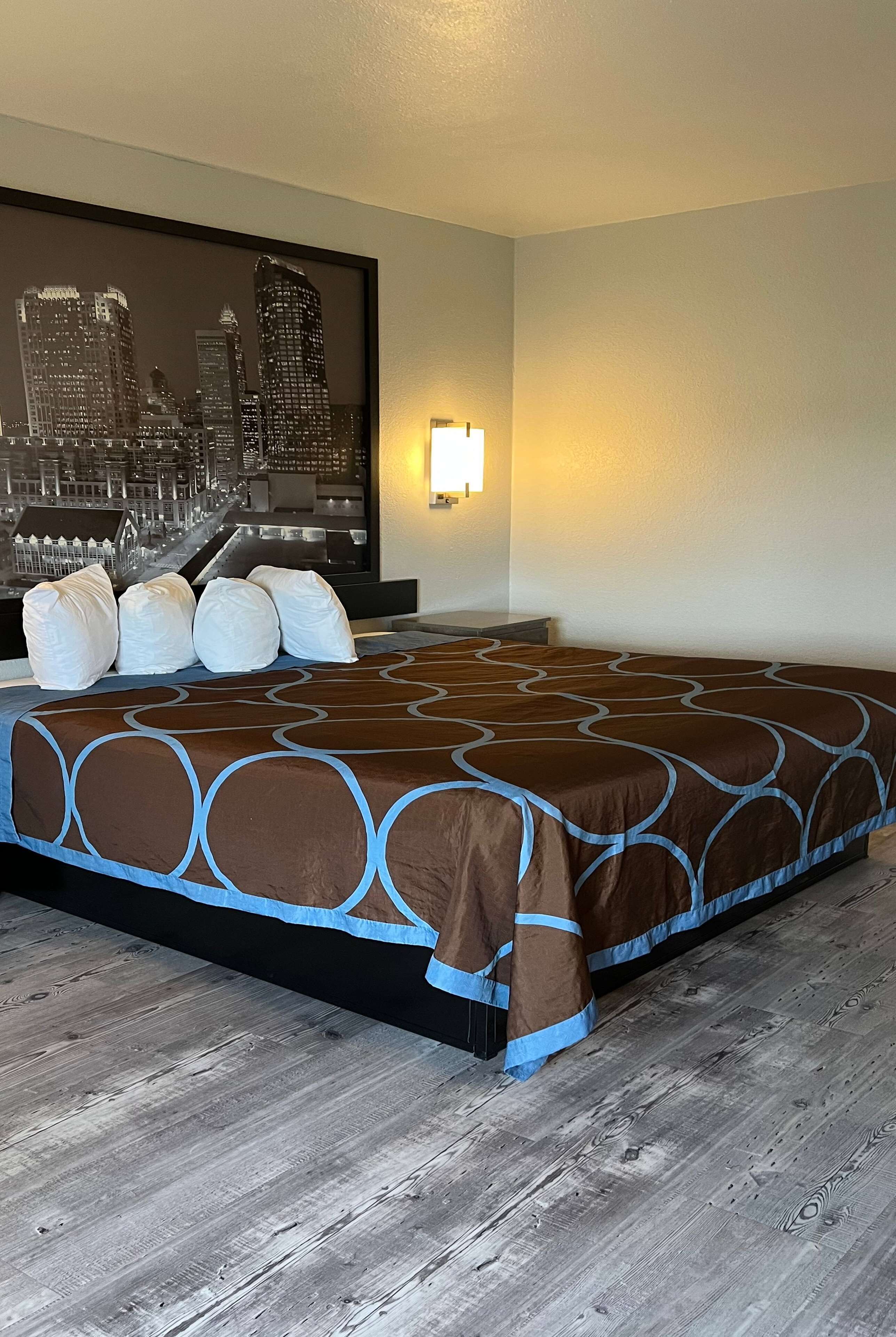 A hotel room with a large bed, a cityscape wall art, and a nightstand with a lamp.