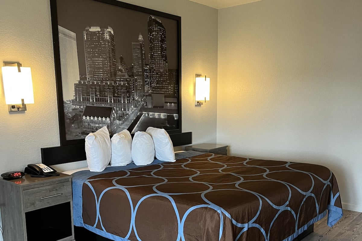 A hotel room with a large bed, a nightstand with a phone, and a cityscape picture on the wall.